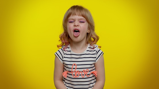 Portrait of cheerful funny blonde child girl 5-6 years old showing tongue making faces at camera, fooling around, joking, aping with silly face, teasing. Yellow studio background. Teen kid children