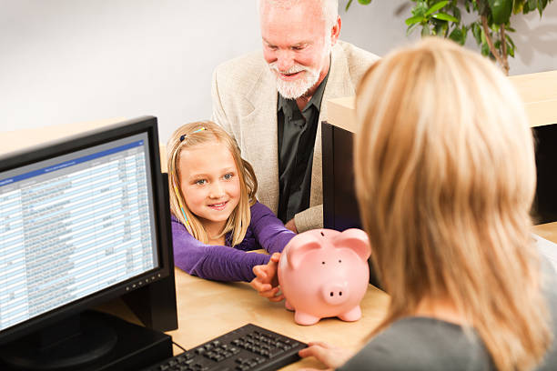 Child Handing Coin Piggy Bank, Opening Bank Account with Teller A cheerful grandparent helping a smiling young girl grandchild with her piggy bank savings. Opening a children bank account with the bank teller in a retail bank counter. Photographed indoors in horizontal format. bank account stock pictures, royalty-free photos & images