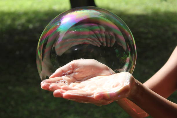 Child holding a bubble in her hands A big soap bubble being held in a girl's hands. Grass and the base of a tree are in the background, out of focus. Taken during the summertime. Landscape photography. bubble wand stock pictures, royalty-free photos & images