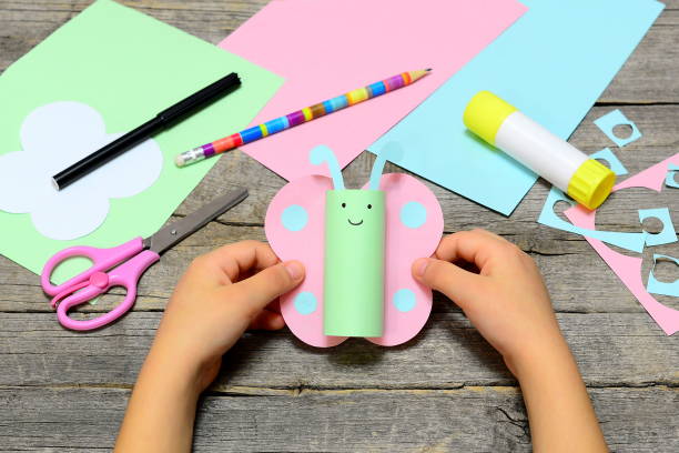 Child holding colored paper butterfly in hands. Child shows a fun paper diy craft. Stationery on an old wooden background. Preschool summer paper art and glue crafts activities. Children creative abilities development stock photo