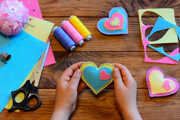 Child holds a felt valentine in his hands. Child made valentines from felt. Valentines day crafts idea. Felt heart ornaments, scissors, thread, felt sheets on a wooden table. Sewing craft projects stock photo