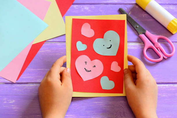 Child is holding a Valentines day card diy in his hands. Small kid made a handmade Valentines day greeting card with hearts smiles. Cute and simple paper crafts for kindergarten. Paper cutting hearts. Closeup. Paper crafts at home. Easy kids paper crafts stock photo