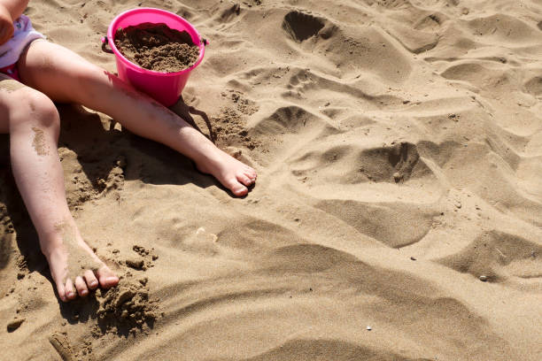 A child plays with a pink plastic bucket in the sand on a beach A child plays with a pink plastic bucket in the sand on a beach. Crete, Greece. Concept for traveling with children / family vacation and holiday. human feet buried in sand. summer beach stock pictures, royalty-free photos & images