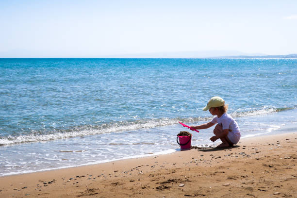 Child plays with sand A child plays with a pink plastic bucket and shovel in the sand on a beach, close to the sea. Crete, Greece. human feet buried in sand. summer beach stock pictures, royalty-free photos & images
