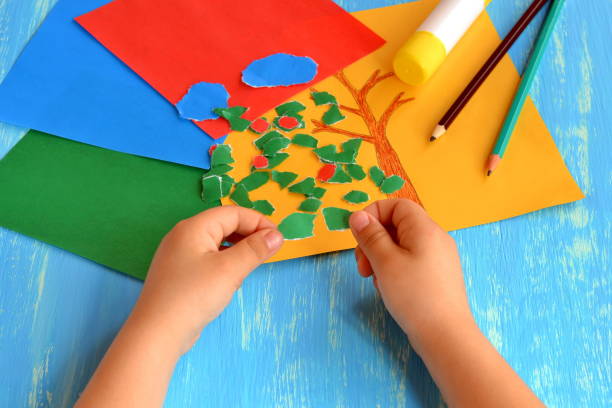 Child tears a red paper into small pieces. Child holds red paper pieces in his hands. Kindergarten art lesson. Set of color paper, pencils, glue stick on wooden background. Fun paper crafts for kids stock photo