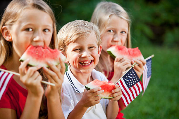 Children at Fourth of July or Memorial Day picnic A group of three children at a picnic on July 4th or Memorial day. They are sitting, eating watermelon and holding small American flags.  They are siblings from the same family, a brother and two older sisters, all with blond hair.  The girls are wearing red shirts and the boy is wearing a white shirt with a blue stripe on his collar.  The focus is on the little boy, who is sitting in the middle with a big grin on his smiling face as he looks at the camera.  He is 4 years old. memorial day stock pictures, royalty-free photos & images