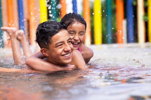 Latino children a teenage boy and an 8-year-old girl play in the pool, cuddle and cuddle in the water while smiling and having fun