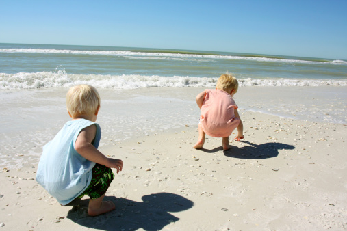 two young children, a boy and his baby brother are on the beach by the ocean shore, picking up seashells