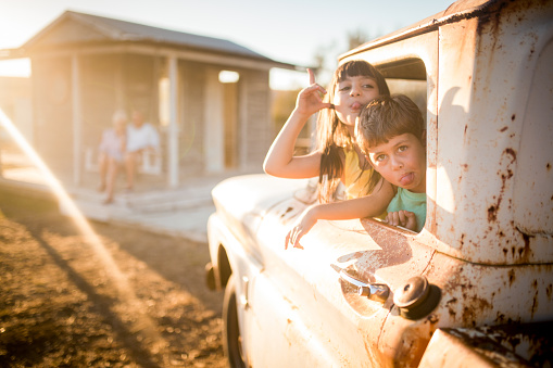Cute children pulling faces from in an old vehicle while their grandparents sit on a porch swing behind