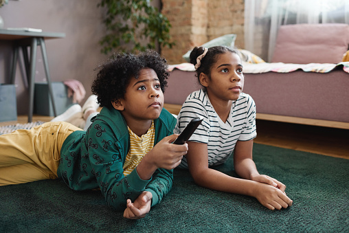 Two African American children lying on floor and watching TV together during leisure time at home