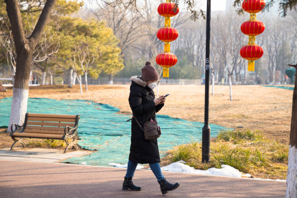 Beijing, China- February, 09, 2020: A Chinese girl wears protective masks is watching her cellphone as having a walk in an empty city park during outbreak of coronavirus（COVID-19） stock photo