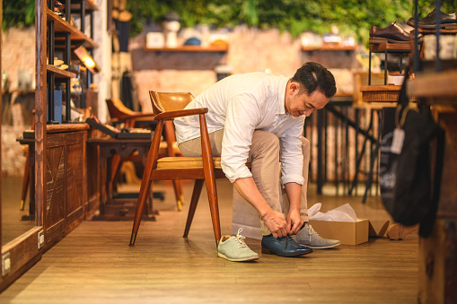 Mid adult Chinese male customer shopping for comfortable natural leather shoes at a retail store. He is sitting on a wooden chair and tying up the shoelaces.  He is happy because he is going to pay a good price for the shoes.