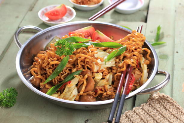 Chinese Style Mie Goreng or Stir Fried Egg Noodles is Traditional Indonesian Food with Chicken, Egg, Tomato, Onion, Vegetable and Sweet Soy Sauce in White Plate Served on Wooden White Table. stock photo
