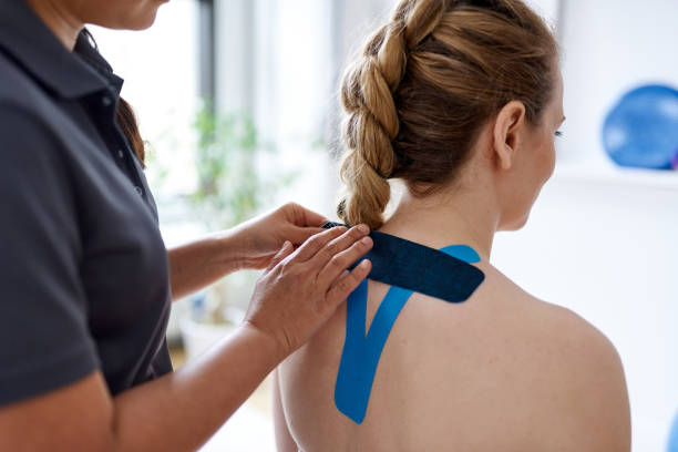 Chinese woman massage therapist applying kinesio tape to the shoulders and neck of an attractive blond client in a bright medical office stock photo