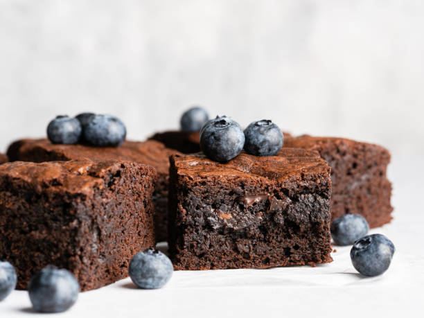 Chocolate brownie squares decorated with fresh blueberry on baking paper. Closeup view, light grey concrete background. Tasty homemade fudge and chewy dessert with melting pieces of chocolate. chewy stock pictures, royalty-free photos & images