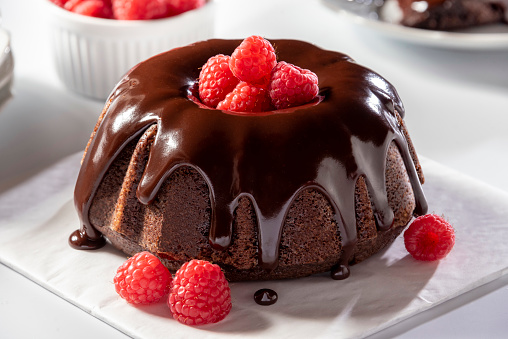 Beautiful mini chocolate bundt cake with chocolate ganache and raspberries. The delicious cake sits on a white table with plates, dish of raspberries and other items in the background out of focus.