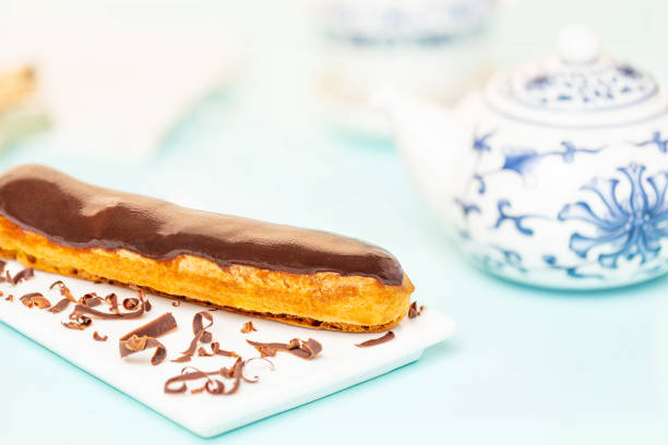 Chocolate French eclair pastry on white plate spread with chocolate shavings, aside Chinese teapot over light blue background. stock photo