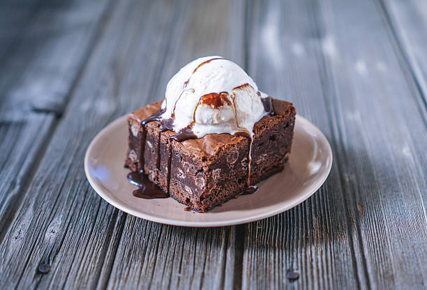 Chocolate Fudgy Brownie with Vanilla Ice Cream on top. Chocolate Fudgy Brownie with Vanilla Ice Cream and Chocolate Syrup on Wood Background. Selective focus, toning. brownie stock pictures, royalty-free photos & images
