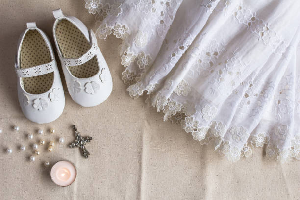 Christening background with baptism dress, shoes, candles and crystal cross pendant on white linen cloth - top view stock photo