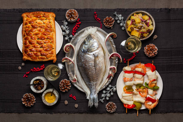 Christmas eve dinner concept. Baked dorado fish, fish pie, potato salad, seafood skewers. White wine glases. Festive decoration. Catholic tradition related to the Feast. Top view. stock photo