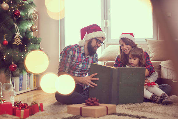 Christmas fairy tale Young parents sitting on the living room floor next to a nicely decorated Christmas tree, mother holding a baby girl in her lap and all together reading a fairy tale christmas story telling stock pictures, royalty-free photos & images