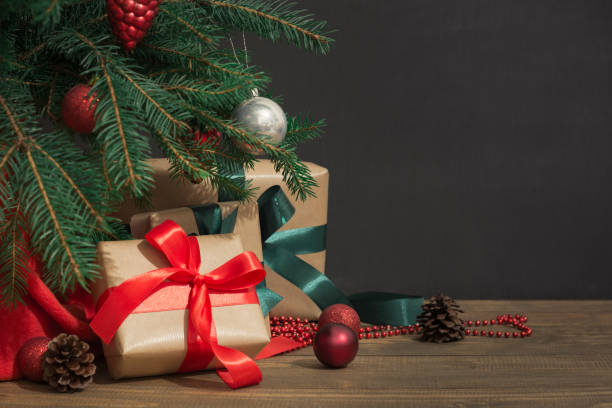 Christmas holiday background. Gifts with a red ribbon, Santa's hat and decor under a Christmas tree on a wooden board. Christmas holiday background. Gifts with a red ribbon, Santa's hat and decor under a Christmas tree on a wooden board. Close up. Copy space on chalkboard. below stock pictures, royalty-free photos & images