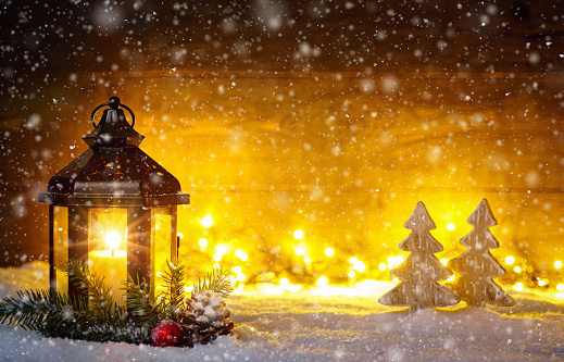 Christmas scene with a lantern, trees, fir branch, snow flakes and blurred lights in front of an illuminated dark wooden board as copy space.