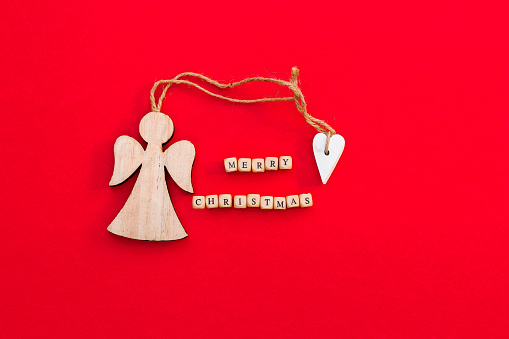 Christmas wooden angel with heart and twine with words Merry Christmas on little bricks on bright red background. Minimalism concept. Natural eco materials New Year toy. Copy space. Top view. Postcard template