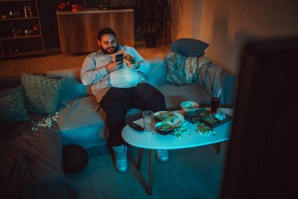 Chubby guy sitting on sofa at home One man, sitting at home, using mobile phone while watching movie. fat man looks at the phone stock pictures, royalty-free photos & images