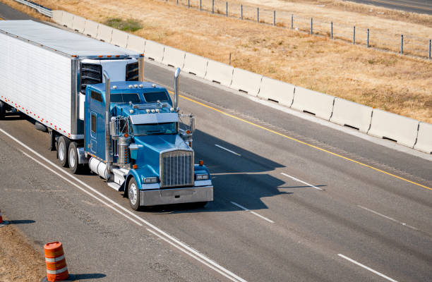 Classic idol blue big rig semi truck with chrome parts and cab with truck driver rest compartment with upper windows transporting cargo in refrigerated semi trailer running on the wide highway road stock photo