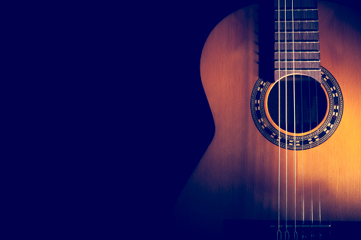 Classical Guitar on a dark background. Guitar wood is light brown with nice grain. Guitar has nylon strings with a pattern around the sound hole.