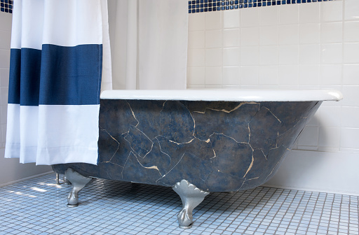 Old-fashioned, cast iron clawfoot tub decorated with torn wallpaper pieces with blue & white shower curtain.