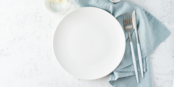 Clean empty white plate, glass of water, fork and knife on white stone table, copy space, mock up, top view, long banner