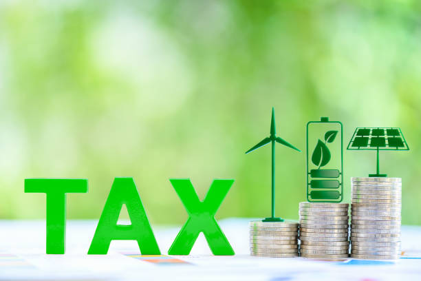 Clean, renewable energy or electricity production tax credits and incentives, financial concept : Green energy symbols atop coin stack e.g solar panel, wind turbine, fuel cell battery and the word TAX stock photo