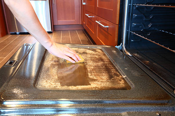 Cleaning dirty greasy oven A close-up image of an open oven glass door with dirty greasy stains on it covered with special chemical foam for oven cleaning and woman's hand holding a cleaning sponge cleaning this oven.  Ceramic tile floor on the background. Modern kitchen interior. Dirty oven. Glass oven door is being cleaned. oven stock pictures, royalty-free photos & images