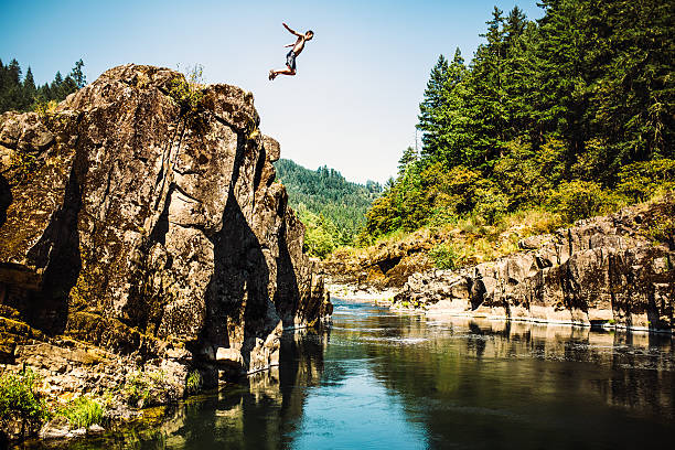Cliff Jumping Man A man jumps from a large ledge over a pristine river, his arms out like he's flying.  Horizontal image. cliff jumping stock pictures, royalty-free photos & images