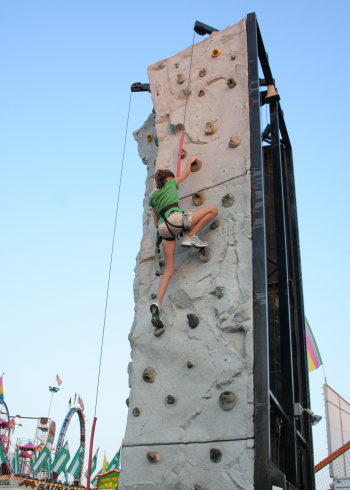 a teenage girl scales a climbing wall at a fair/carnival. various rides in the lower background give context.  setting is near dusk.