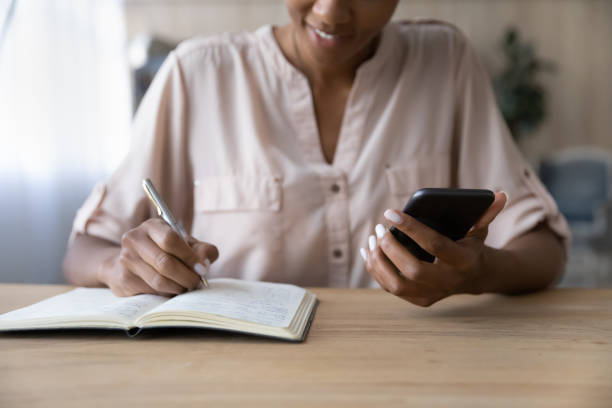 Close up African American woman holding phone, taking notes Close up African American woman holding phone, taking notes in notebook, checking, writing down important information, businesswoman planning workday, student watching webinar, studying online Exam prep apps stock pictures, royalty-free photos & images