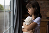 Close up lonely little girl hugging toy, looking out window, standing at home alone, upset unhappy child waiting for parents, thinking about problems, bad relationship in family, psychological trauma