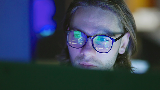 Close-up of a face of a young, long haired man, working at night in a dark office