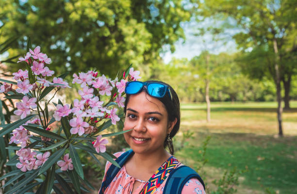 Close up of a beautiful Indian smiling girl standing close to a plant of pink Cherry blossom flower in a butterfly park in Gurugram, Haryana, India. Girl solo tourist in a garden on a sunny morning. A simple Indian college student enjoying in Nature and witnessing the beautiful flowers in spring time at a garden in the city. A much needed change from the busy life of the city. haryana stock pictures, royalty-free photos & images