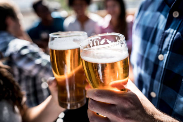 Close up of a customers at a bar holding a beer and making a toast Close up of a customers at a bar holding a beer and making a toast with people having fun at the background artisanal food and drink photos stock pictures, royalty-free photos & images