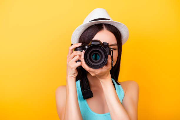 nahaufnahme eines fotografen machen einen schuss auf eine digitale kamera in urlaub. sie trägt sommer casual-outfit und einen hut auf leuchtend gelbem hintergrund - bauen fotos stock-fotos und bilder