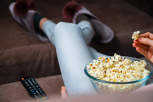 Close up of hand waking popcorn from a bowl while watching TV. Person sitting in comfortable couch and watching home cinema in the dark.