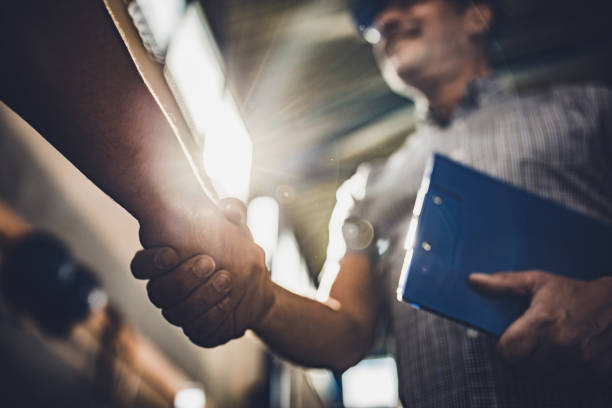 Close up of inspector shaking hands with unrecognizable person in a factory. Low angle view of manager greeting unrecognizable person in industrial building. blue collar worker stock pictures, royalty-free photos & images