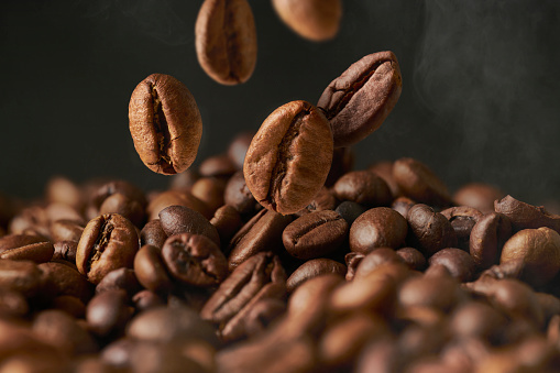 Close up of roasted coffee beans on a black smokey background. The grains fall from above in a group.