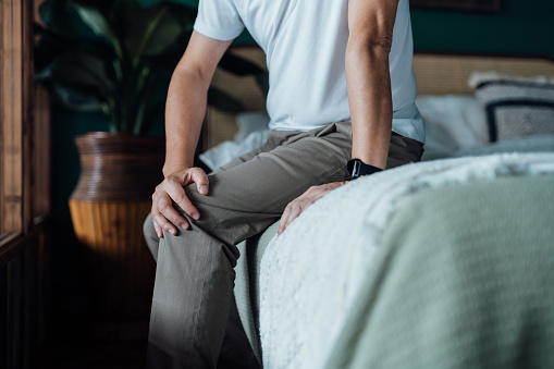 Close up of senior man holding his knee in discomfort, suffering from knee pain while sitting on bed at home. Elderly and health issues concept