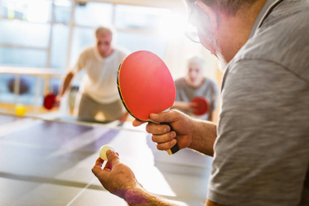 Close up of senior man playing table tennis in health club. Close up of senior man serving while playing table tennis with friends in a health club. table tennis stock pictures, royalty-free photos & images
