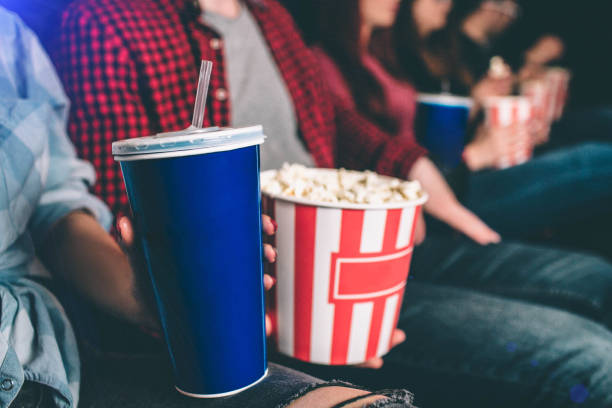 Close up of tasty but unhealthy food. There are basket of popcorn and a blue cup of coke on picture. Man and woman are holding it together. stock photo