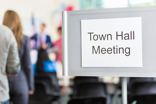 Group of people gather for town hall meeting. A 'Town Hall Meeting' sign is at the entrance of the meeting room. The crowd is blurred in the background.
